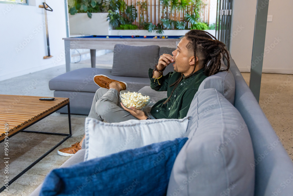 Obraz premium African American male sitting on gray sectional holding bowl of popcorn in lounge near pool table