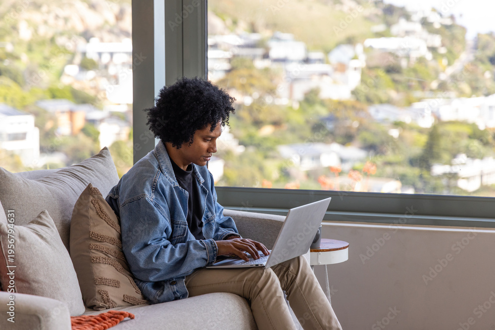 Fototapeta premium African American adult man sitting on beige sofa at home wearing denim jacket, using laptop