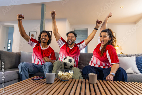 Diverse friends in red white jerseys cheering on grey sectional sofa at home, holding soccer ball