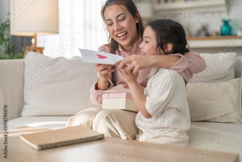 Asian happy mother reading handmade greeting card from little daughter on sofa, Cute child giving gift box to mom, Mother's Day celebration, sweet family bonding, childcare and warm love life concept