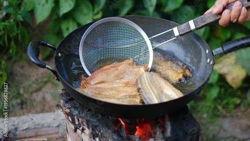 Cooking Fresh Fish in a Traditional Pan Over Open Fire with Vegetation in the Background and Cooking Tools in the Scene