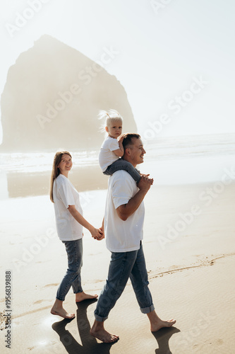The image shows a family walking along a sandy beach by the sea. The man carries a small child on his shoulders, supporting him with his arms. The child sits quietly and looks forward.