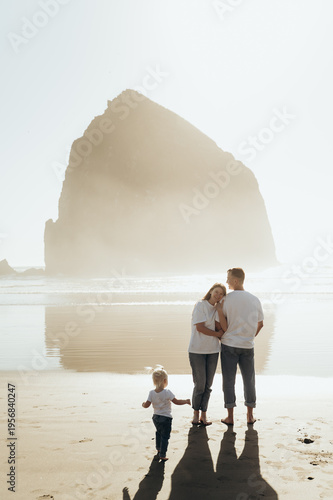 he photograph depicts a family standing on the seashore. The man and woman embrace, creating an atmosphere of warmth, love, and support. A small child stands next to them, clinging to their parents, f