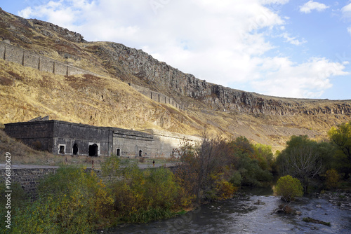 Historical stone buildings sit embedded in a rugged hillside overlooking a winding river and autumn foliage near Kars, Turkey.