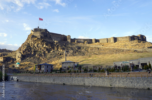 Massive black basalt stone walls and defensive ramparts of the Kars Citadel rise prominently above the surrounding hills and modern urban landscape