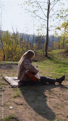 Blonde woman playing string guitar outdoors in autumn forest. Concept of sound therapy, mental health and wellness rituals. Calmness tranquility audio-sensory practices. Aura farming energy holistic