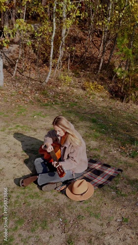 Beautiful blonde girl playing acoustic guitar and singing relaxing enjoying outdoors on sunny autumn day. Hippie woman in boho style learning and practicing playing musical instrument. Fall leaves