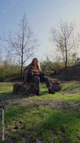 Blonde woman playing string guitar outdoors in autumn forest. Concept of sound therapy, mental health and wellness rituals. Calmness tranquility audio-sensory practices. Aura farming energy holistic