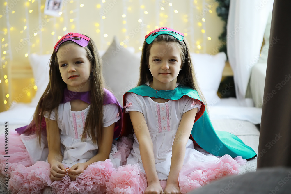 Fototapeta premium Twin Girls in Princess Costumes with Capes and Masks Sitting on Bed with Fairy Lights