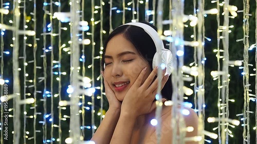 Close up of a beautiful young Asian woman wearing white wireless headphones, listening to music with her eyes closed. Dreamy atmosphere surrounded by glowing fairy lights bokeh.
