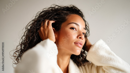 Young woman in white towel robe applying hair mask mousse to wet curly dark hair after shower, home hair care styling treatment, natural light, wellness lifestyle concept