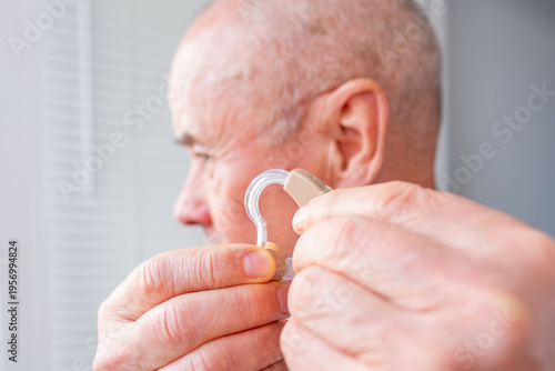 Senior man holding behind-the-ear hearing aid in hand, adjusting beige BTE, focus on modern device, medical solution hearing loss, deafness elderly people, hearing assistance, medical equipment