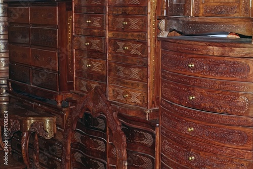 Collection of intricately carved wooden furniture is densely arranged, showcasing drawers, cabinets, and decorative patterns with detailed craftsmanship.