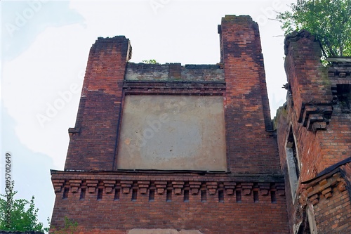 Weathered brick wall with missing sections stands against the sky, evoking a sense of decay and history. Vegetation grows atop, highlighting nature's reclaim.