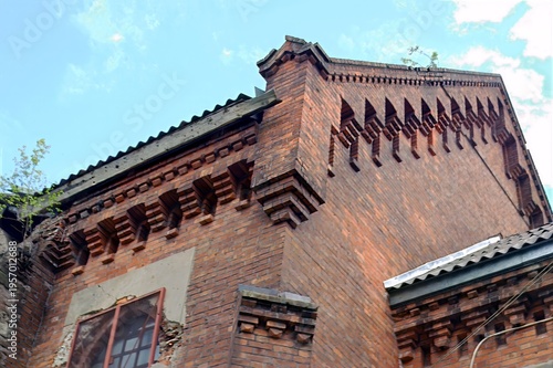 Old brick building facade displaying intricate geometric patterns, with minor vegetation growth and a clear sky in the background.