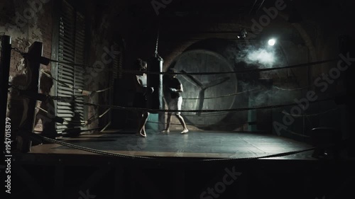 Box fighters training on dark ring in fitness club, sport workout in fight club. Young adult african man and woman practicing arm strikes on punching bag, healthy lifestyle and sport activity for body