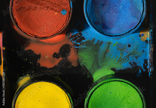 Top view of cracked watercolor pans in red, blue, yellow, and green, surrounded by vivid paint splashes. Dramatic lighting enhances texture, creating an abstract, energetic, and artistic composition.