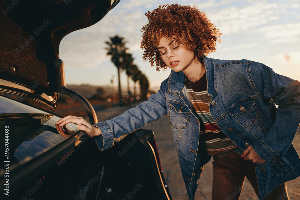 Fototapeta premium Young woman with curly hair wearing rainbow sweater and denim jacket, smiling while opening car trunk outdoors, casual lifestyle, warm sunlight, relaxed mood, natural background