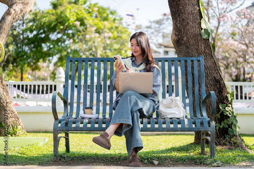 Φωτογραφία Pretty asian woman looking at phone over laptop while sitting cross legged on bench in garden park