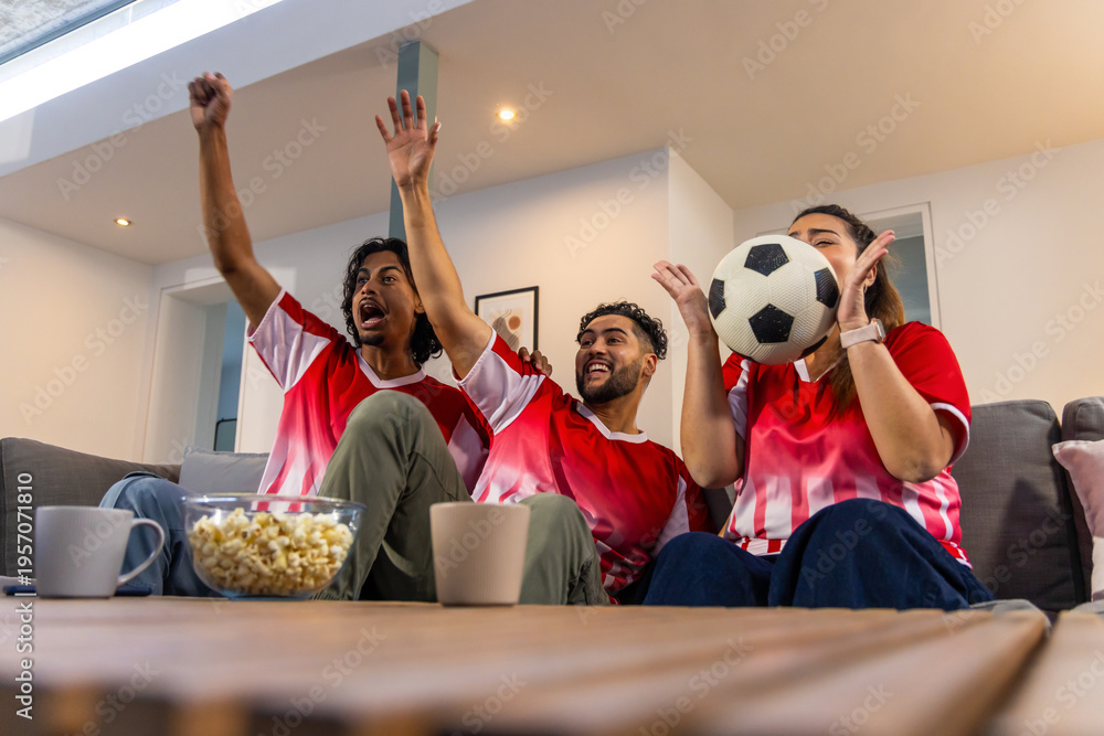 Fototapeta premium Friends wearing red white jerseys cheering, watching match on sofa with soccer ball, popcorn, mugs