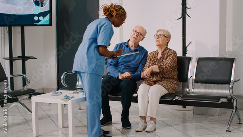 African american nurse talking to senior people in waiting room, waiting to attend medical consultation with doctor. Assistant filling in checkup report files before examination appointment.