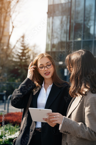 Women discussing business ideas while using a tablet in an outdoor office setting near city buildings during daylight hours
