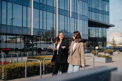 Women in business attire walk together outside a modern office building while discussing work and using a tablet in a city setting during the day