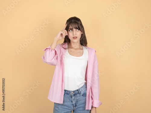 Thoughtful young woman touching her temple and looking aside, wearing pink shirt and denim, standing against beige studio background with copy space. Thinking and decision concept.