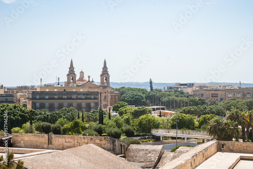 Stone walls, pedestrian bridge and trees of Biskuttin Gardens, buildings and towers of St. Publius Parish Church in the background, Valletta MALTA