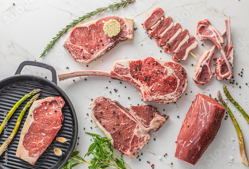 Top view of various premium raw beef cuts including Tomahawk, T-Bone, ribeye, and lamb chops on white marble, with a cast iron grill pan, asparagus, and spices, concept for butcher shop and gourmet co