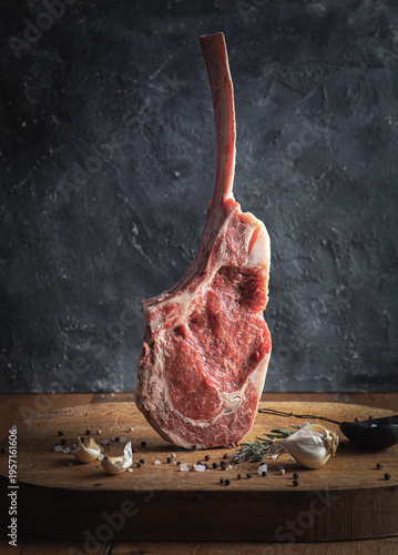 Raw Tomahawk ribeye steak standing on a wooden board with garlic, rosemary, and spices. Gourmet bone-in beef on a dark textured background.