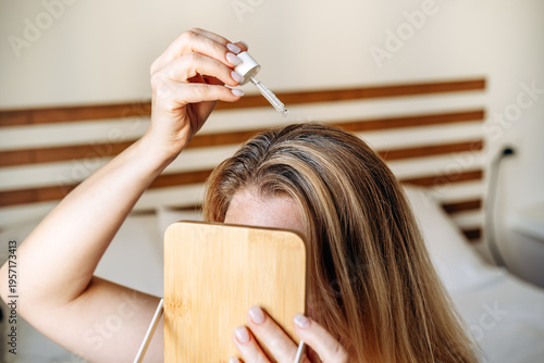 Woman applying hair serum with dropper to scalp while lying on a bed, focusing on hair care and beauty routine