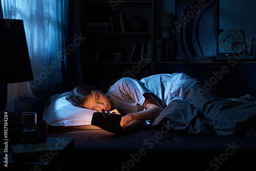 Obraz na plátně Portrait of teenage girl lying in bed at night holding smartphone close to face,