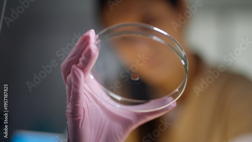 Female scientist in protective gear working in a modern laboratory to test DNA samples. Holding a small glass petri dish with a blood sample up to the camera. Selective focus
