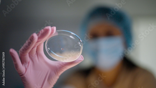 Female scientist in protective gear working in a modern laboratory to test DNA samples. Holding a small glass petri dish with a blood sample up to the camera. Selective focus