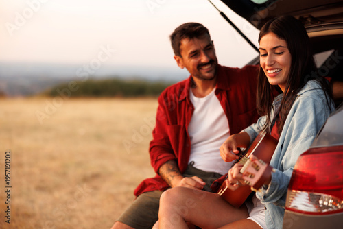 Couple having fun sitting in a car trunk singing and relaxing, woman playing the guitar