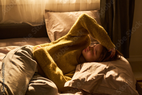 Bild auf Leinwand Young woman lying on bed holding head with hand, appearing to experience discomf