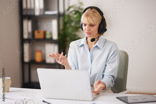A support agent is providing assistance to customers while using a laptop and wearing a headset. The office is organized with shelves and plants in the background.