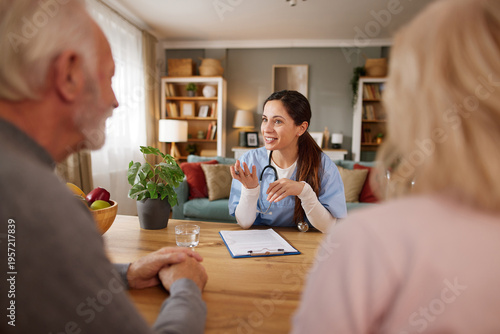 Home health nurse speaks with elderly couple at their home. They sit at a table with notes and a drink while discussing care plans and health needs.