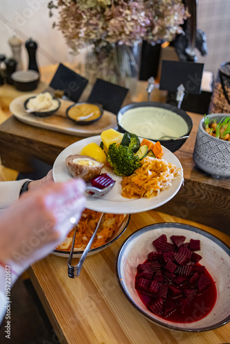 Hand holding a plate filled with steamed vegetables, shredded salad, and a stuffed egg at a buffet station with sauces and beetroot salad visible. Background softly blurred