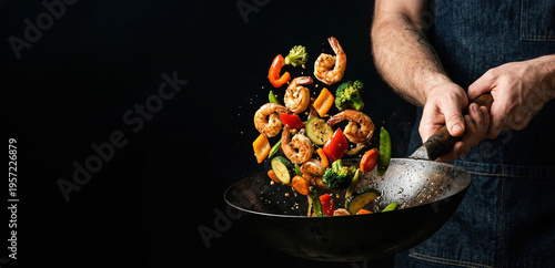 A chef stands in a dark kitchen and uses a wok to stir fry shrimp and colorful vegetables. The food is lifted in the air in a lively motion, showing action in cooking