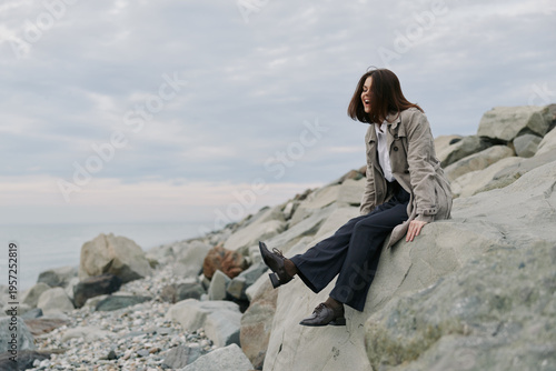 Young woman sits on rocky shore wearing casual clothes, looking away and smiling. Overcast sky creates calm atmosphere by the sea. Concept of relaxation and nature escape.