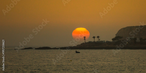 The setting sun descends over the Atlantic Ocean towards the cliffs as people play in the sea and on the beach at the end of another hot day in Taghazout, Agadir, Morocco