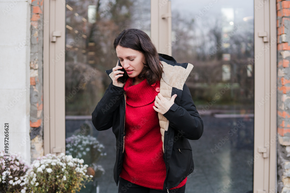 Fototapeta premium Caucasian woman in her 40s with dark hair wearing warm clothes talking on the phone outdoors during her working time. Mature female managing her routine or personal tasks and breathing fresh air.