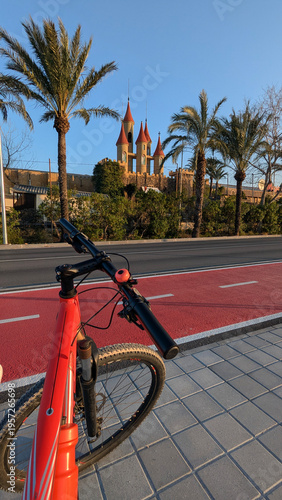POV cycling on a red bike lane in Alicante, Spain, bright red mountain bike handlebars, palm trees, and fairylike castle building background under clear blue sky, active urban travel