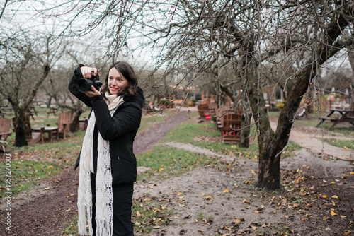Caucasian woman photographer in her 40s wearing warm clothes taking photos of her friend in autumn park. Relaxed mature females spending time outdoors, enjoying leisure time, walking and posing.