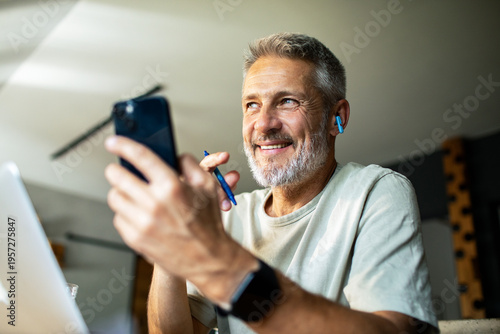 Smiling mature man on smartphone with earbuds at home office