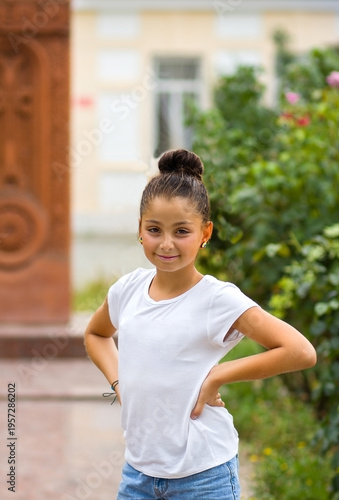 A full-length portrait of a cute teenage girl in a white T-shirt. She is standing in front of a building and green plants, her hands on her hips