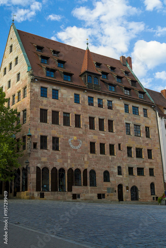 Historic sandstone building in Nurnberg square under blue sky. Steep red roof and dormers frame a tower with a golden star. Arched floor and cobblestone plaza suggest medieval civic pride.