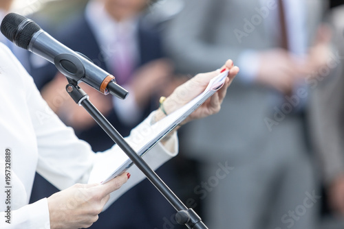 Businesswoman delivering speech at microphone, reading notes during corporate conference or seminar presentation with blurred audience background